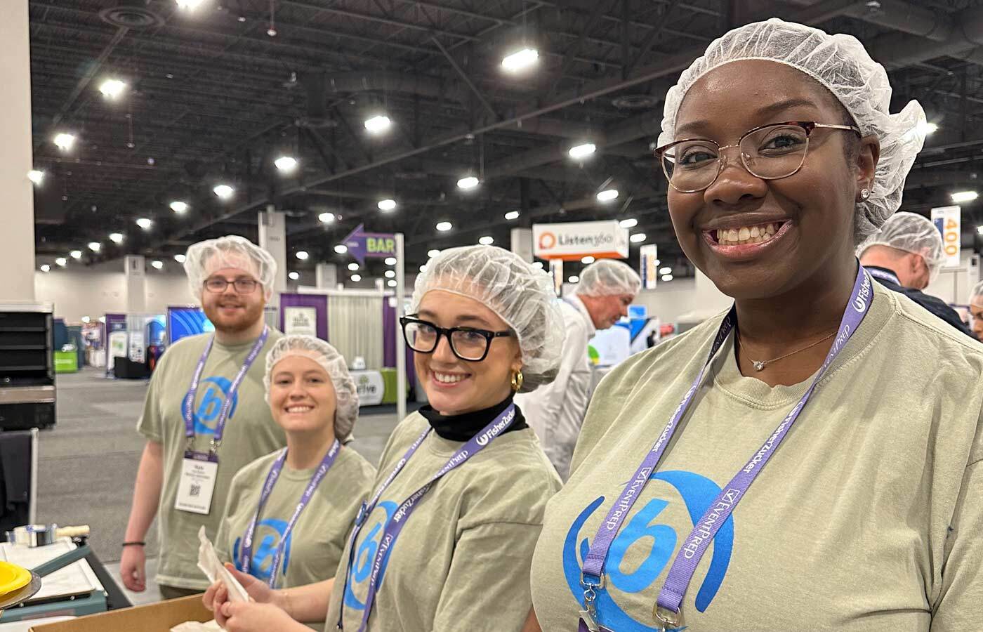 Workers in hairnets in a expo type place standing in front of a folding table.